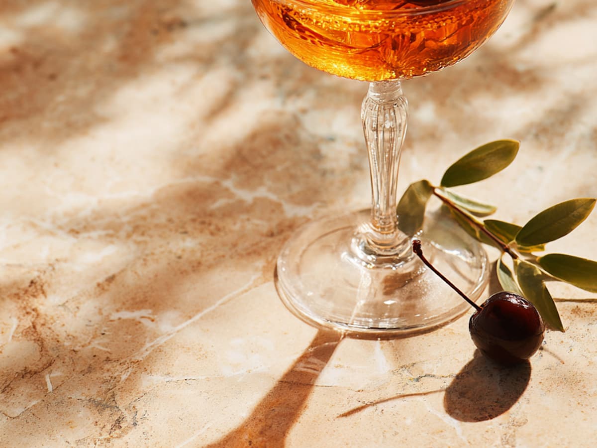 An amber cocktail served in a coupe glass with cherries, resting on the marble bar top.