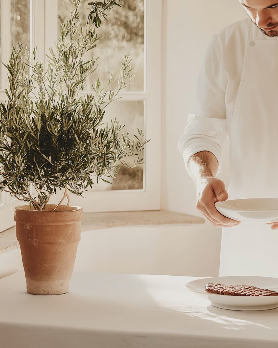 Marco Bello in white chef’s whites, holding a mixing bowl beside a plate of grilled meat and a potted olive tree.